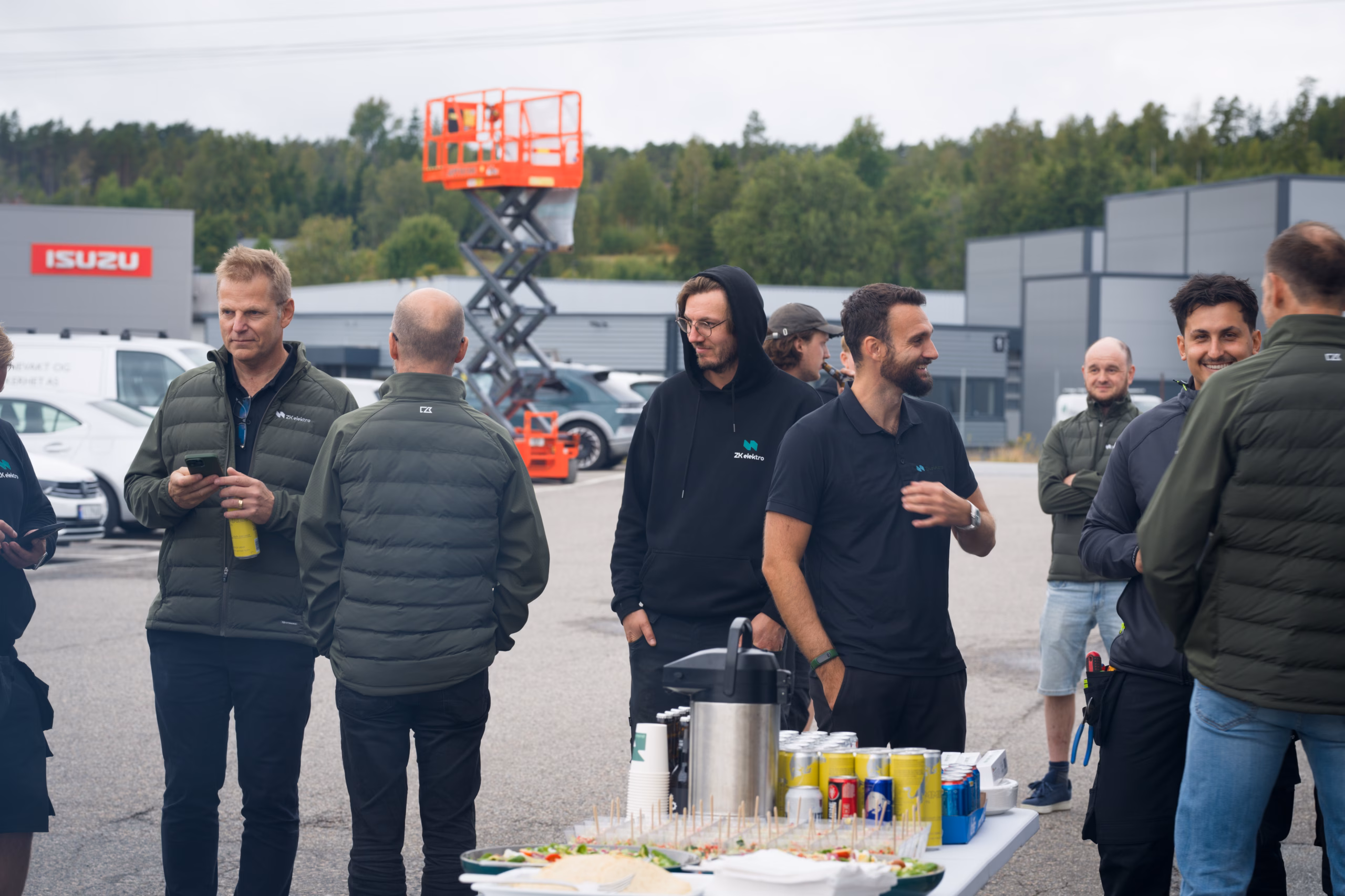 Kollegaer står og snakker ved et bord med mat og drikke under en utendørs samling.